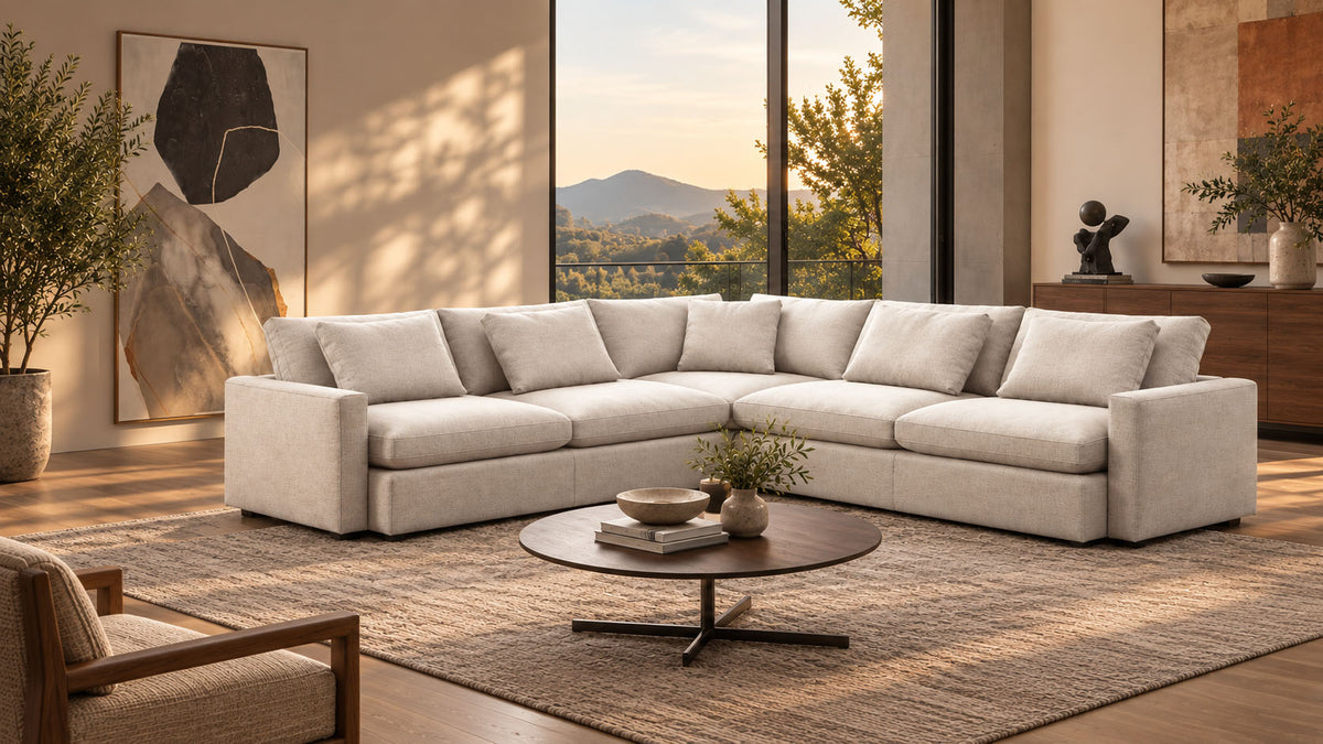 A modern sectional sofa in a light beige or off-white fabric, arranged in an L-shape in a stylishly decorated living room with a round coffee table, textured rug, and large windows showing a sunset view.